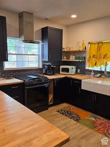 Kitchen featuring dark cabinetry, wood-finish countertops, stainless steel chimney-style range hood, subway tile backsplash, and white apron-front sink