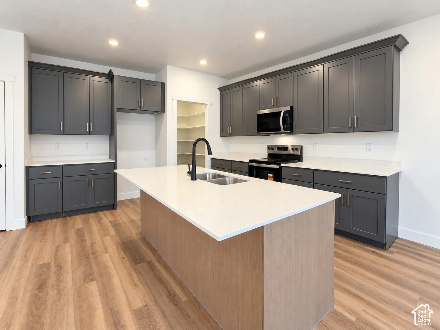 Kitchen featuring dark cabinetry, expansive white countertops, a center island with a dual-basin sink and matte black faucet, integrated stainless steel appliances, and wood-finish flooring