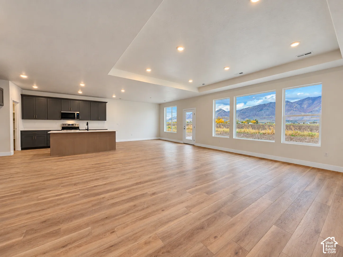 Open-concept interior featuring wood-finish flooring, tray ceiling with recessed lighting, dark-toned cabinetry, kitchen island, and large windows with mountain scenery