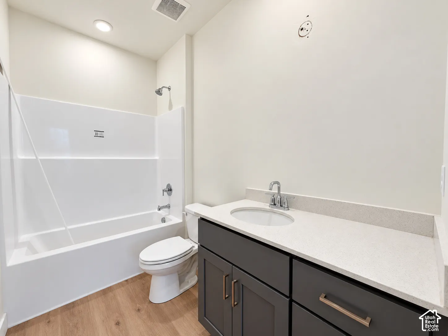 Bathroom featuring wood-finish flooring, dark-toned cabinetry, light-colored solid surface countertop with an integrated sink, and a white acrylic tub-shower combination