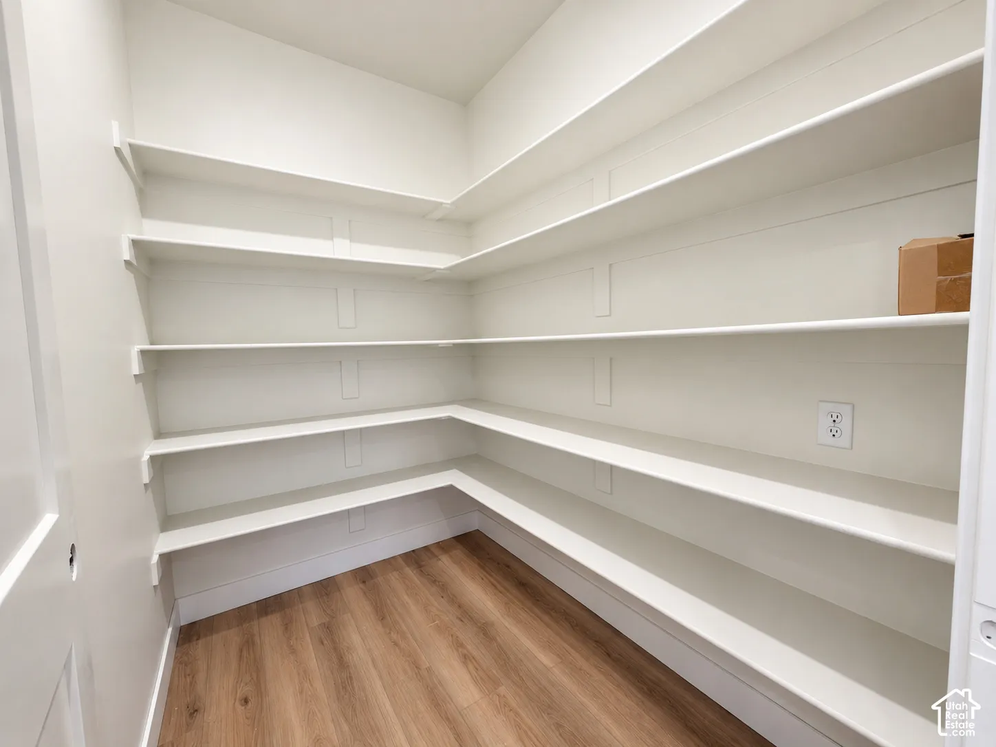 Storage space featuring wrap-around fixed shelving, white painted wall finish, and wood-finish flooring with an integrated electrical outlet