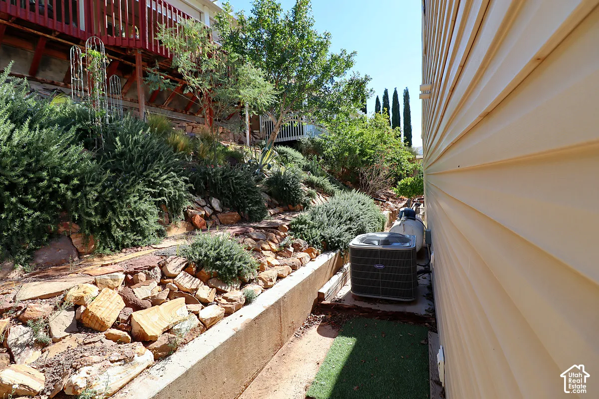 Side yard landscape featuring tiered concrete retaining walls, natural stone ground cover, mature shrubbery, and an exterior HVAC unit
