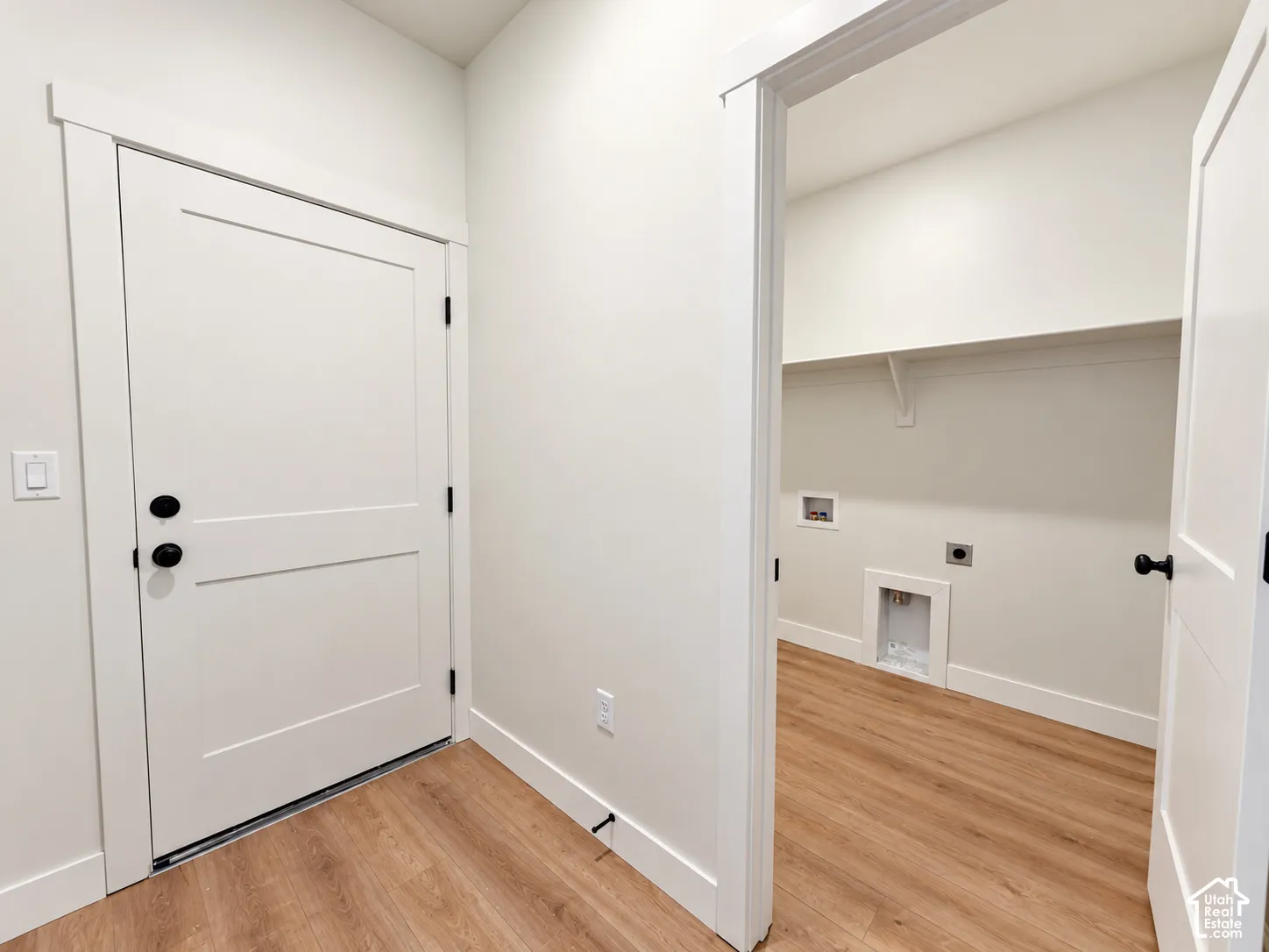Utility area featuring wood-finish flooring, white shaker-style doors with matte black hardware, and an overhead storage shelf