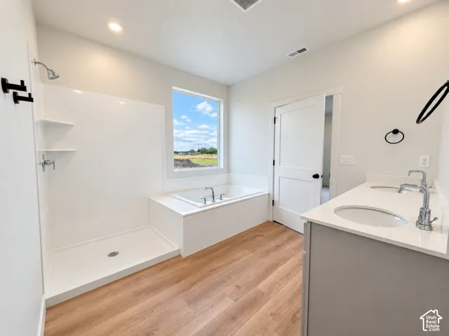 Double vanity featuring a light-toned countertop, wood-finish flooring, a deep soaking tub, and a separate walk-in shower with white surround panels