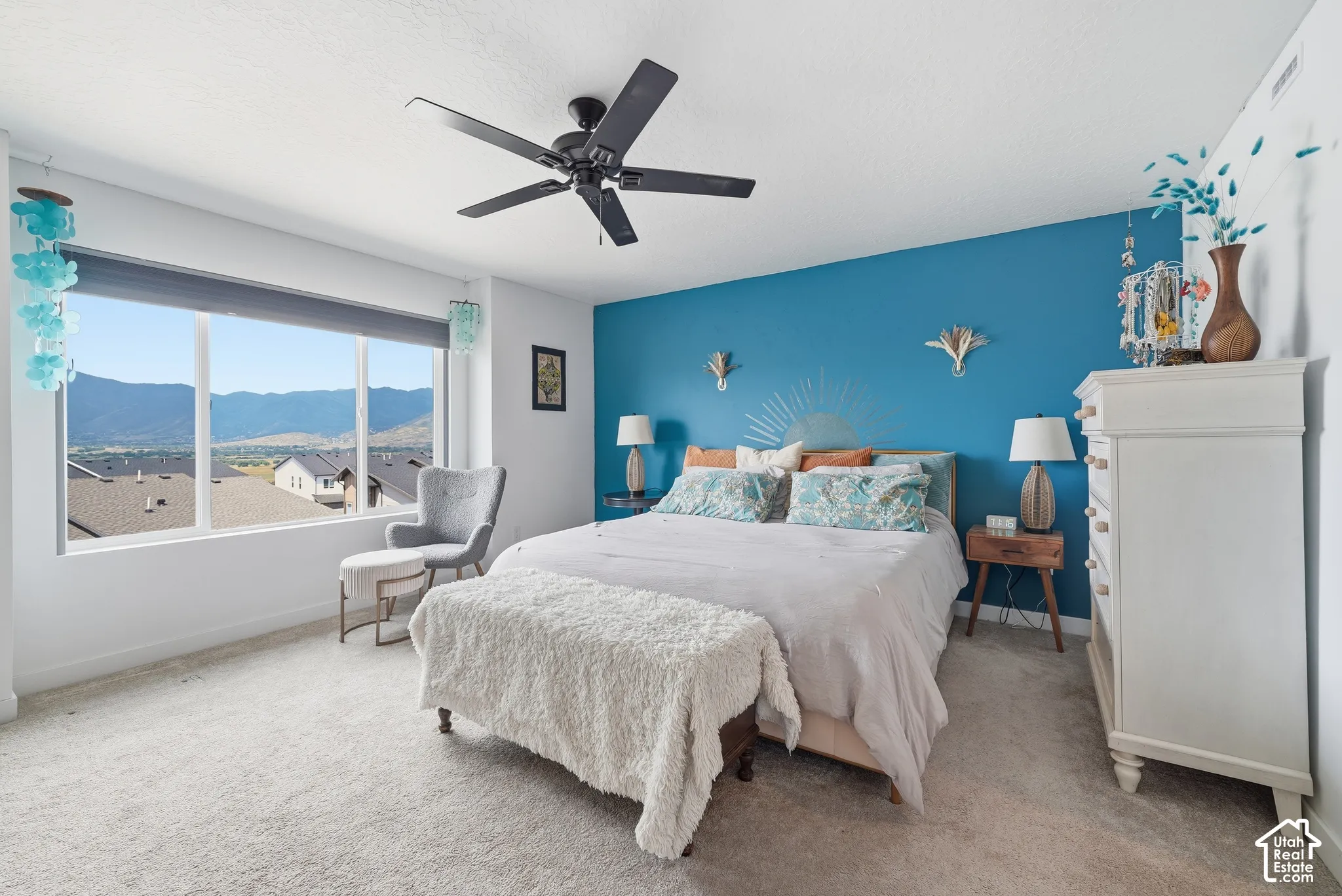 Bedroom featuring mountain backdrop scenery, large picture window, bold accent wall, neutral carpeting, and ceiling fan