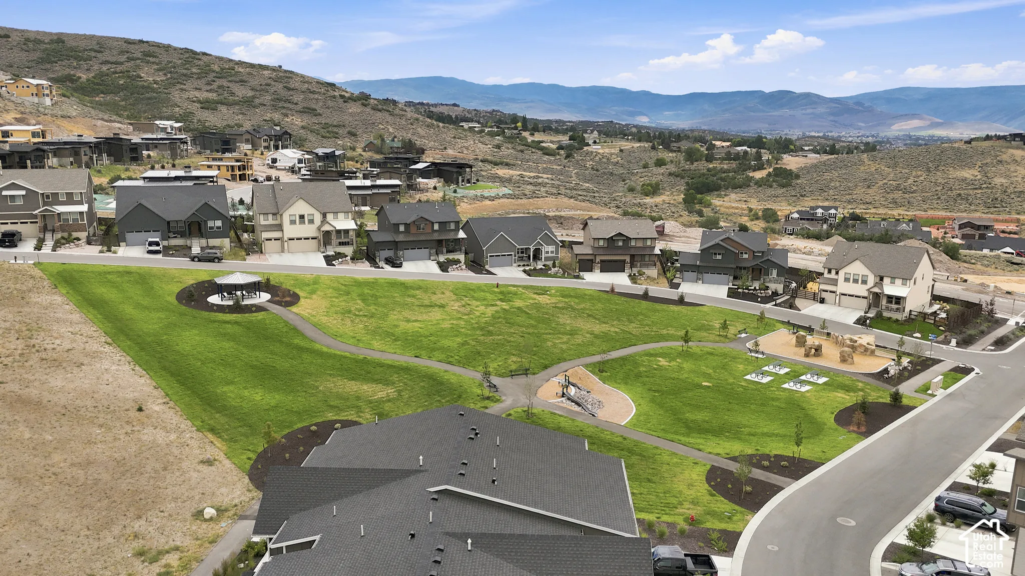 Expansive community green space featuring manicured lawns, paved walking paths, a shaded gazebo, and a mountain-range backdrop