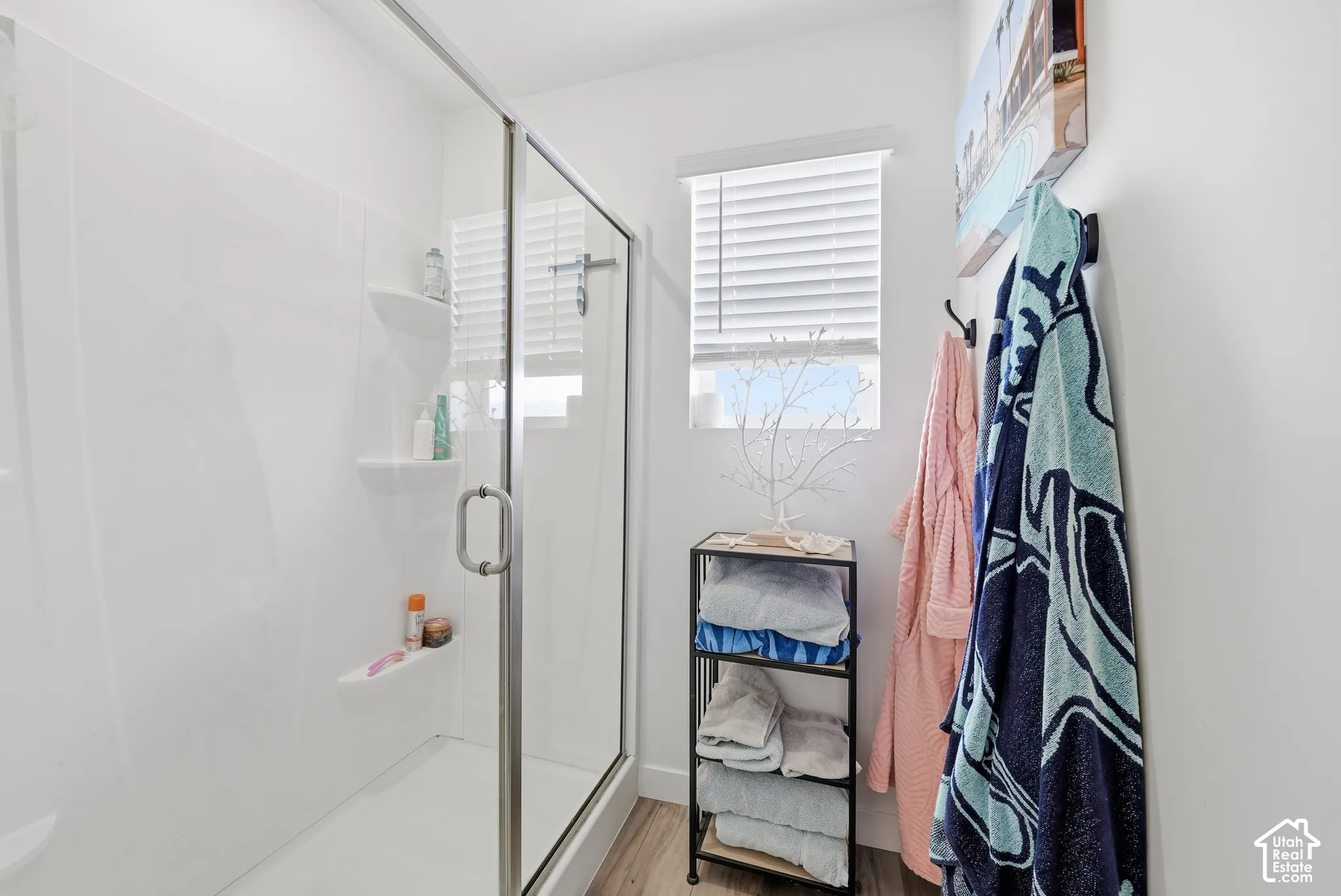 Walk-in shower with white surround, integrated shelving, glass enclosure, window with horizontal blinds, and wood-finish flooring