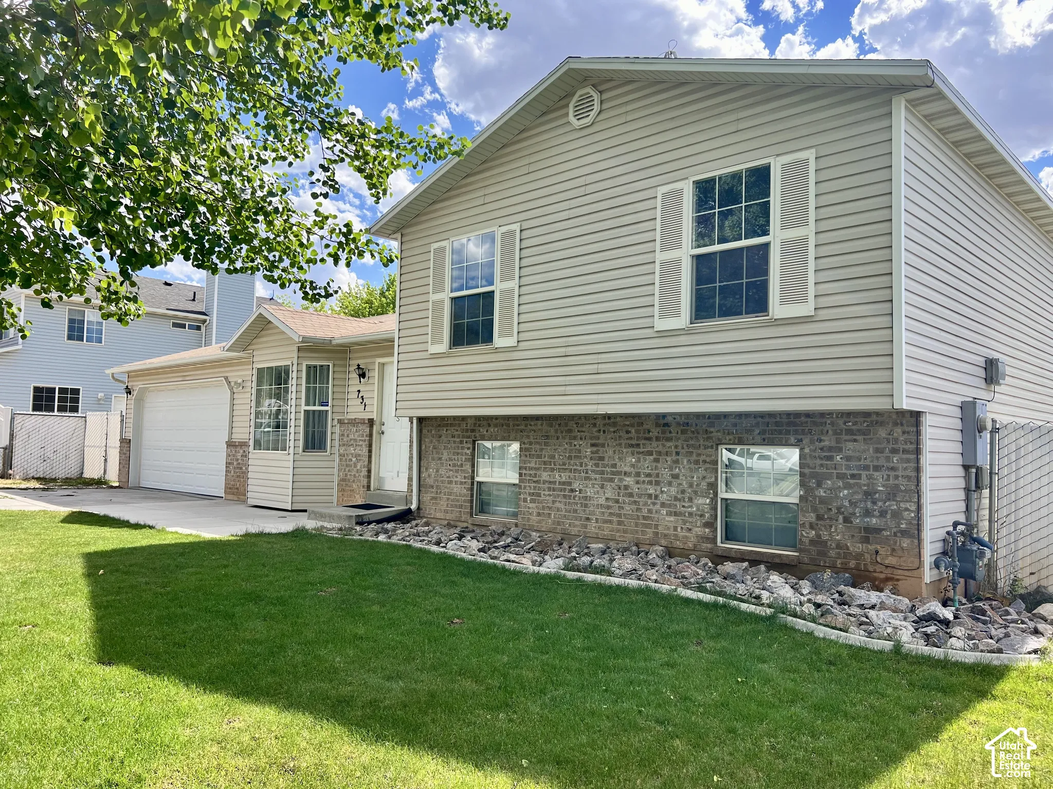 Split-level exterior featuring light beige siding, brick masonry, shutter-trimmed windows, a concrete driveway, and a front lawn with a rock garden border