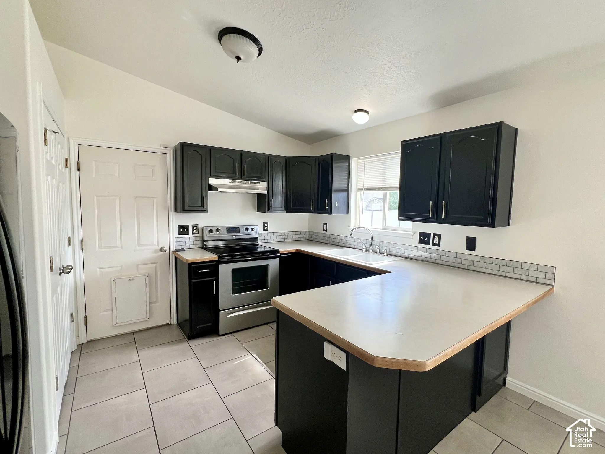 Kitchen featuring dark cabinetry, stainless steel range with overhead hood, light-toned countertops, subway tile backsplash, and ceramic tile flooring