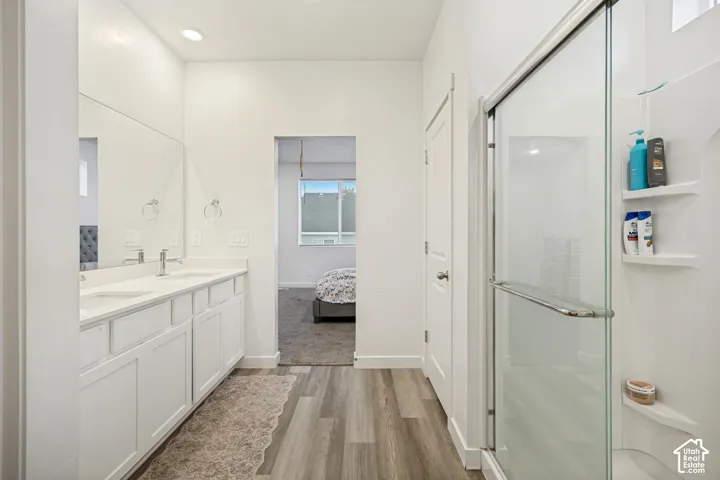 Bathroom featuring a dual-sink vanity with white cabinetry, wood-finish flooring, full-width mirror, and a glass-enclosed shower