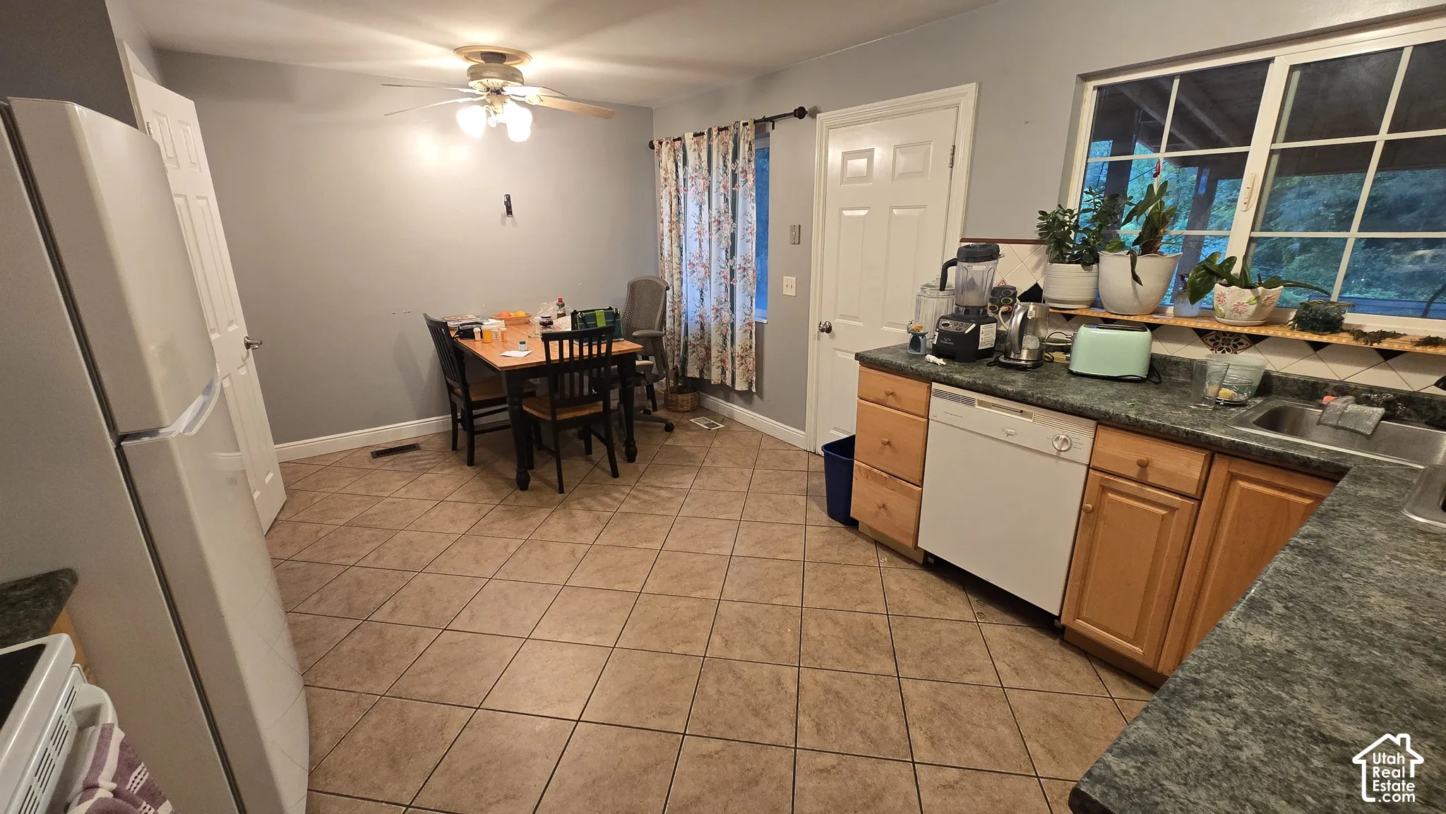 Kitchen area featuring tile flooring, wood-finish cabinetry, stone-texture laminate countertops, a built-in dishwasher, and a ceiling-mounted light fixture with fan