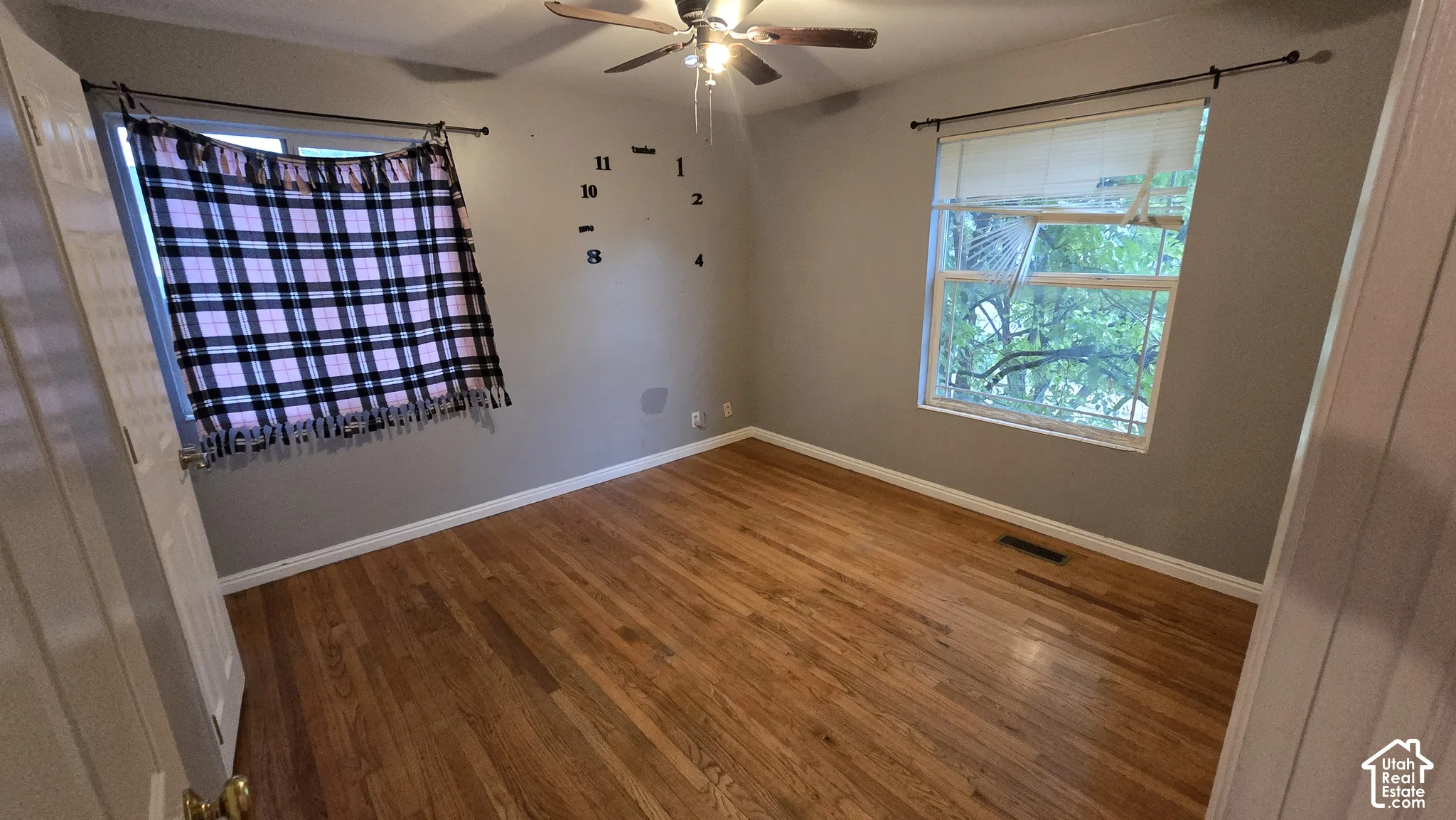 Room featuring wood-finish flooring, dual windows with curtain rods, and a ceiling-mounted light fixture with fan
