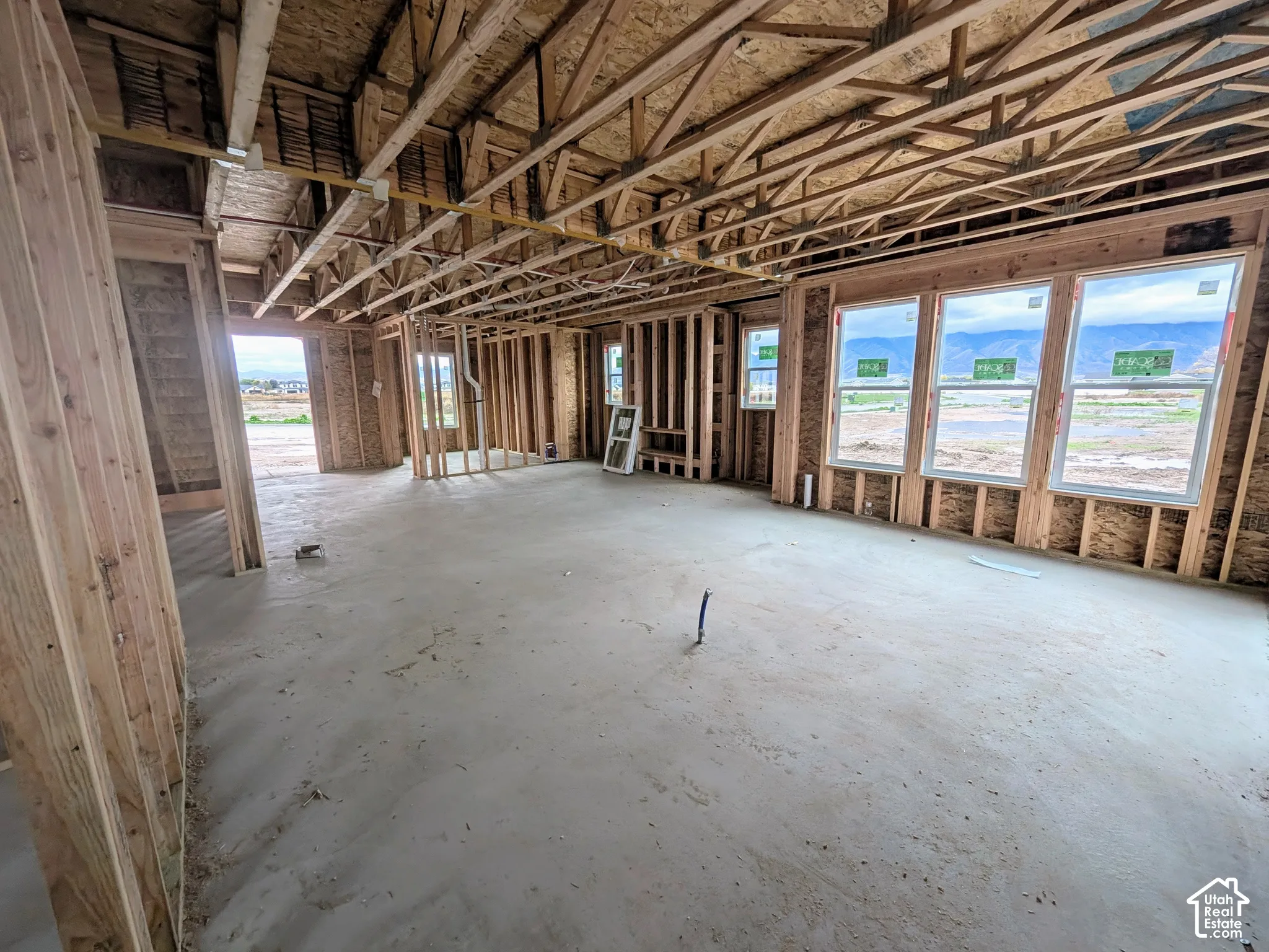 Interior framing featuring exposed wood stud walls, open-truss ceiling rafters, concrete slab flooring, and triple-pane window installations with mountain silhouettes