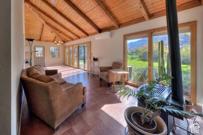 Vaulted ceiling with exposed wood beams, ceramic tile flooring, floor-to-ceiling windows, wall-mounted ductless climate control unit, and freestanding metal stove