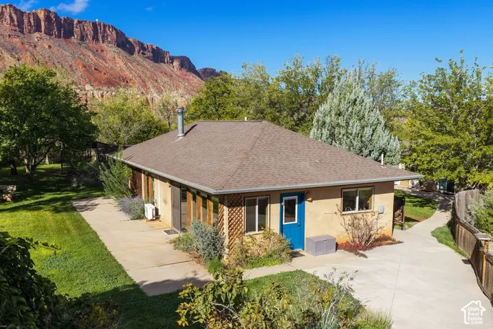 Single-story exterior featuring a stucco facade, hipped roof, concrete patio, and concrete walkway, set against a backdrop of prominent rock formations