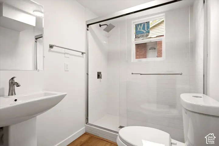 Bathroom featuring a white pedestal sink with a metallic faucet, a walk-in shower with white tile surround and sliding glass doors, a rainfall showerhead, and wood-finish flooring