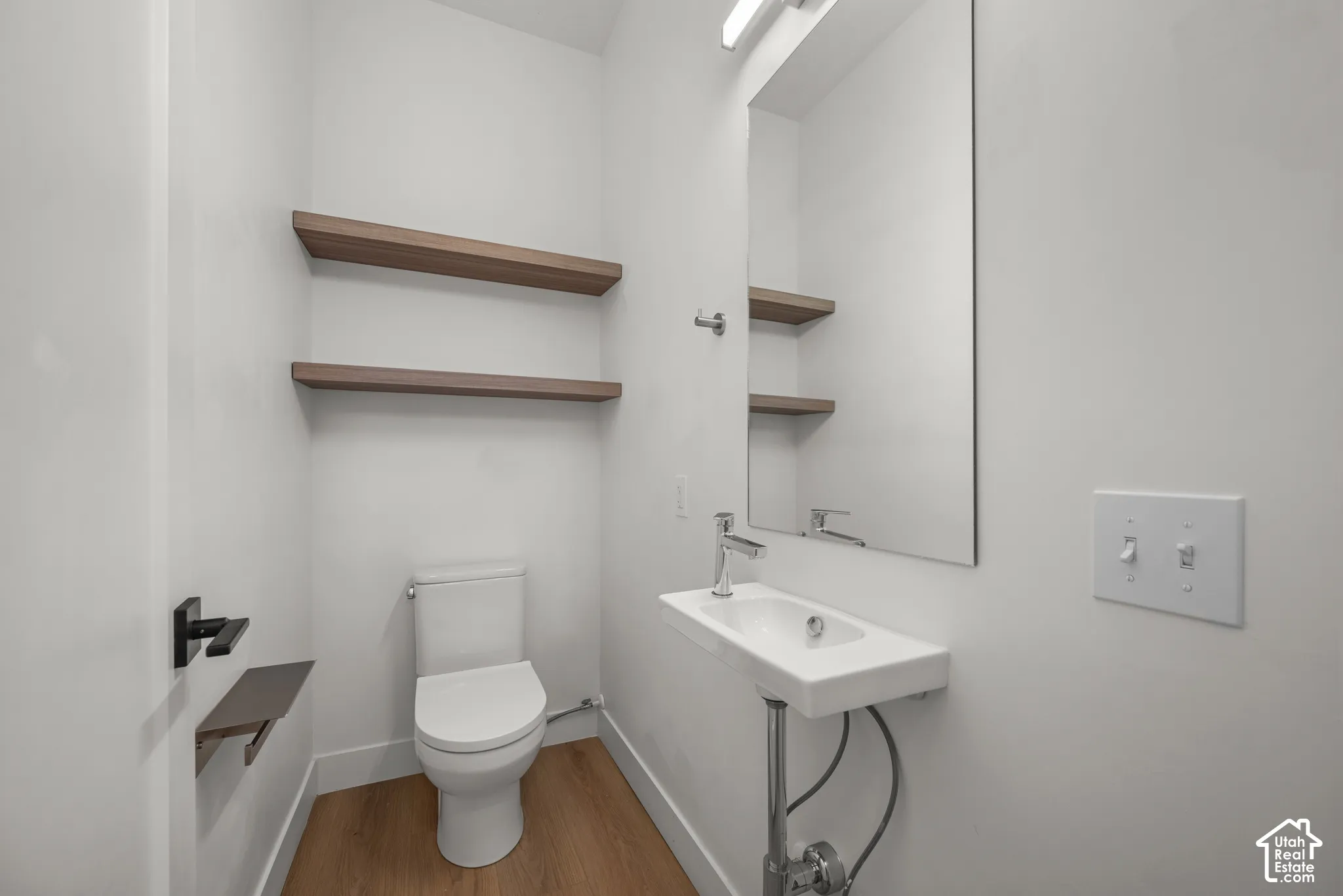 Wall-mounted washbasin with a polished chrome faucet, wood-finish flooring, floating wood-tone shelving, and a frameless wall mirror