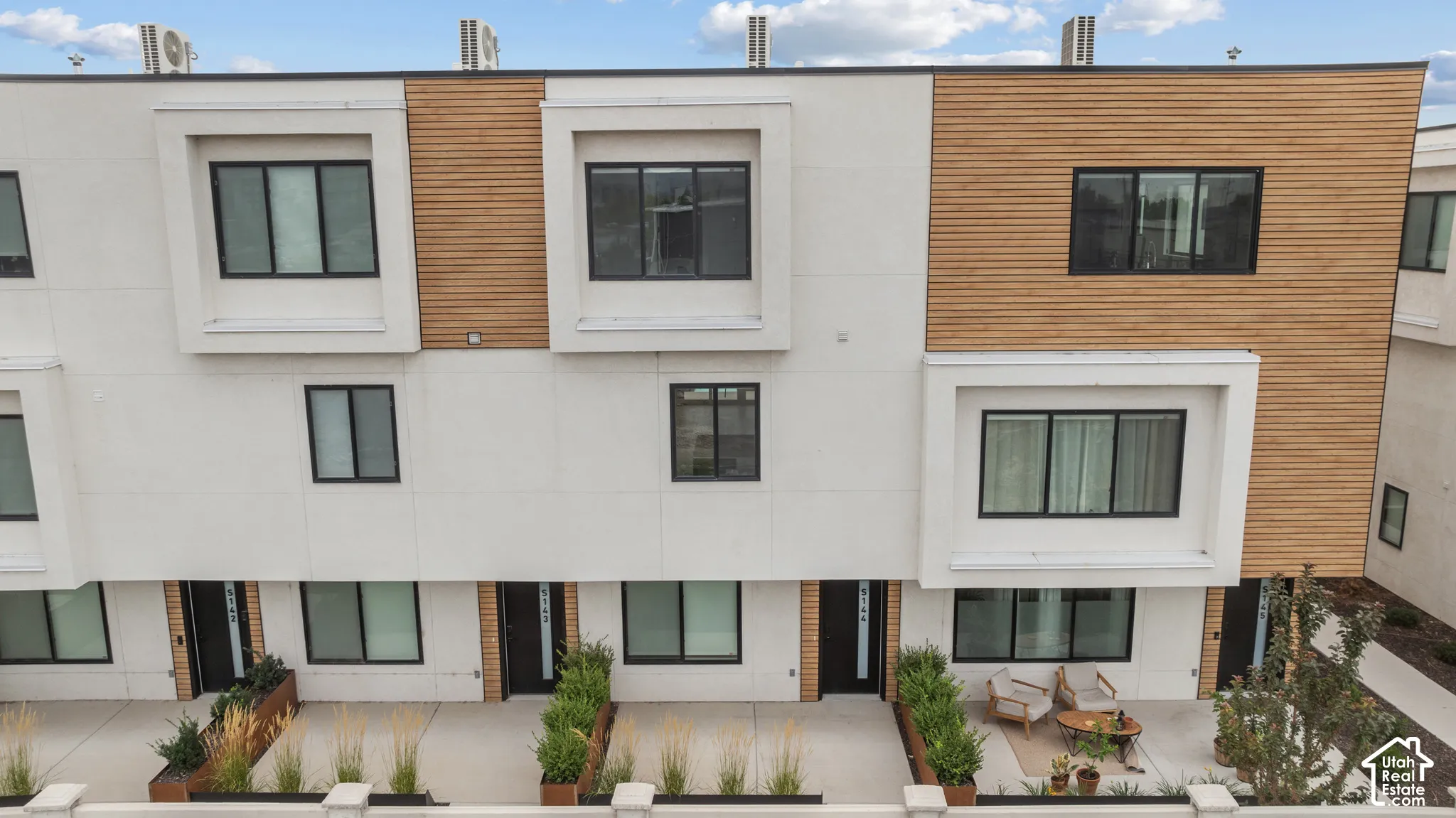 Contemporary multi-level exterior featuring white smooth-panel siding, horizontal wood-slat accents, recessed window bays, and private concrete patios with built-in metal planters