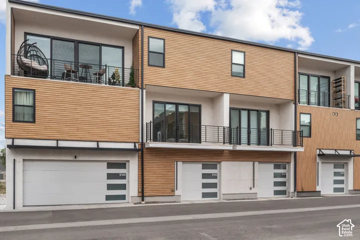 Contemporary multi-story exterior featuring horizontal wood-tone siding, contrasting white concrete lower levels, private balconies with metal railings, and multi-panel garage doors with integrated window panes