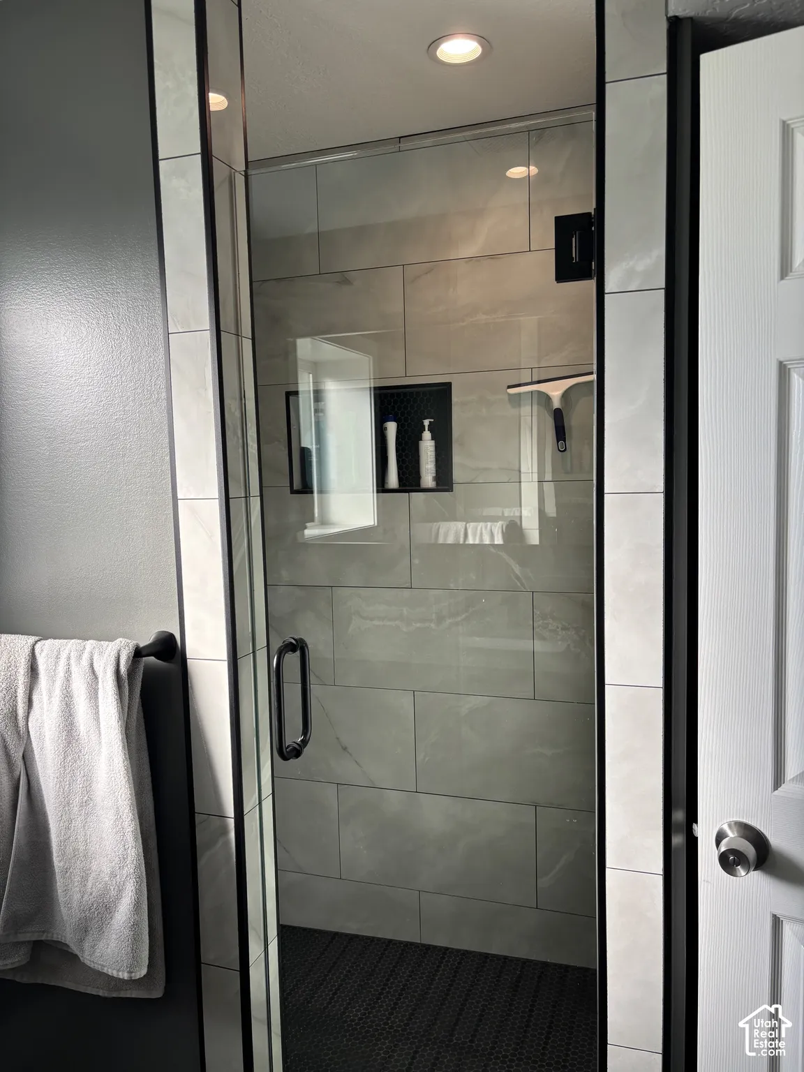Walk-in shower featuring large-format porcelain wall tile, a built-in recessed wall niche, glass door with matte black hardware, and dark mosaic floor tile