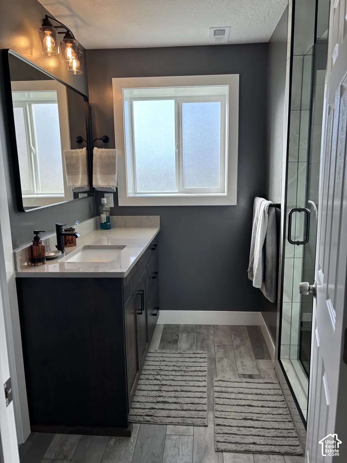 Bathroom featuring a dark wood-finish vanity with a stone countertop, a rectangular under-mount sink, matte black fixtures, and a glass-enclosed shower with tiled walls