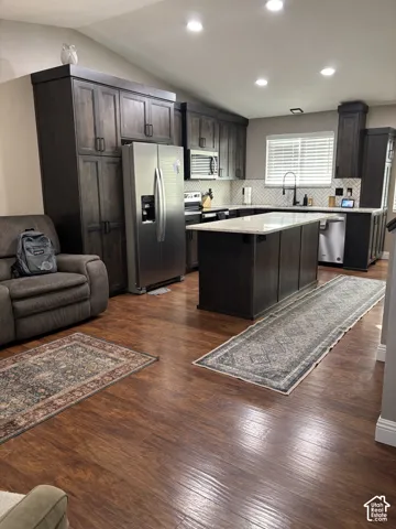 Kitchen featuring dark wood cabinetry, a central island with stone-finish countertops, stainless steel appliances, herringbone-patterned tile backsplash, and wood-finish flooring