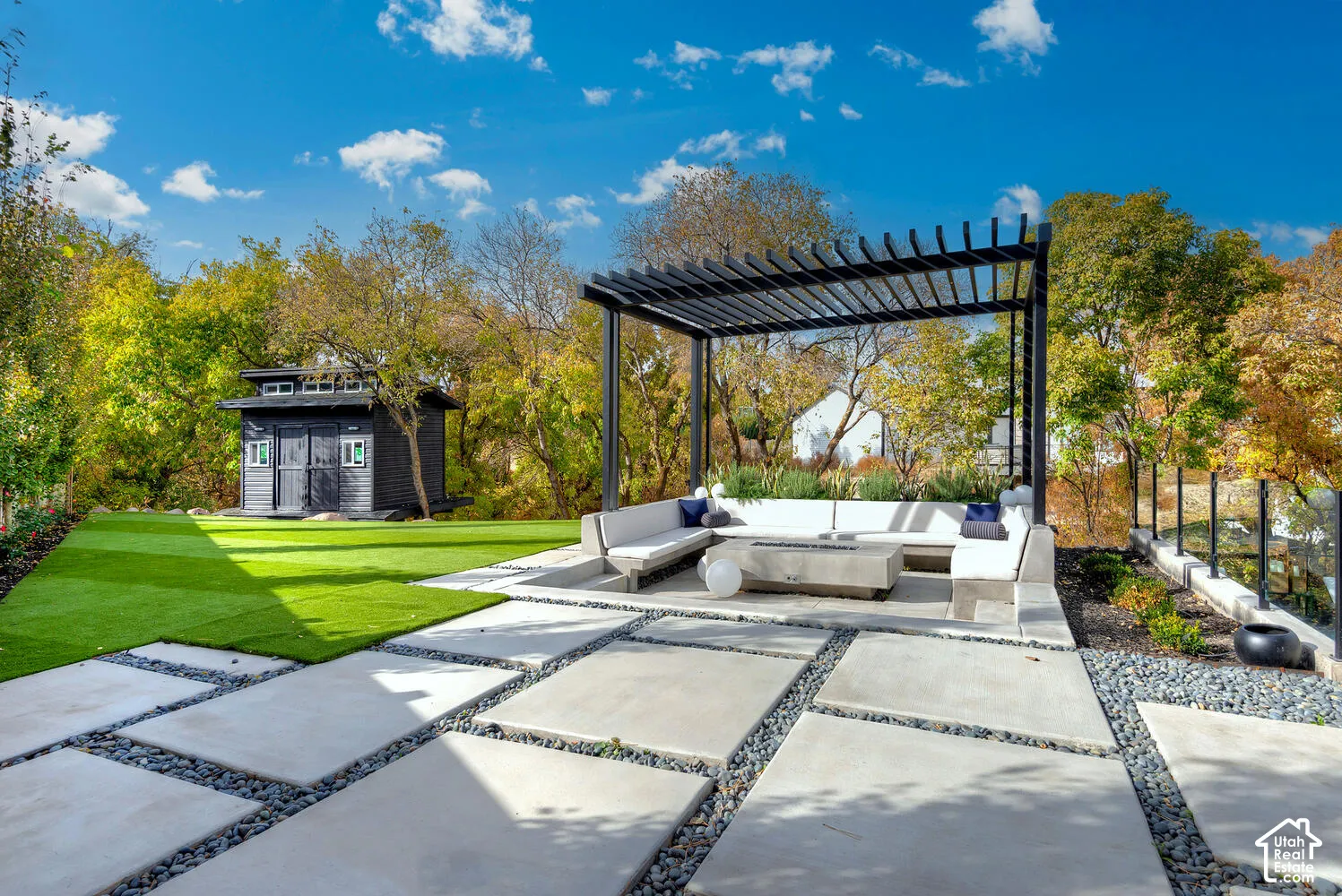Landscaped backyard featuring large-format concrete pavers, artificial turf, a metal pergola, a built-in concrete fire feature, and a detached structure with siding
