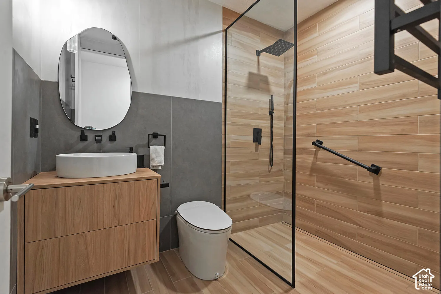 Floating wood-finish vanity with a white vessel sink, charcoal-toned wall tiling, wood-finish tile flooring, walk-in shower with a glass enclosure and matte black fixtures, and a wall-mounted mirror