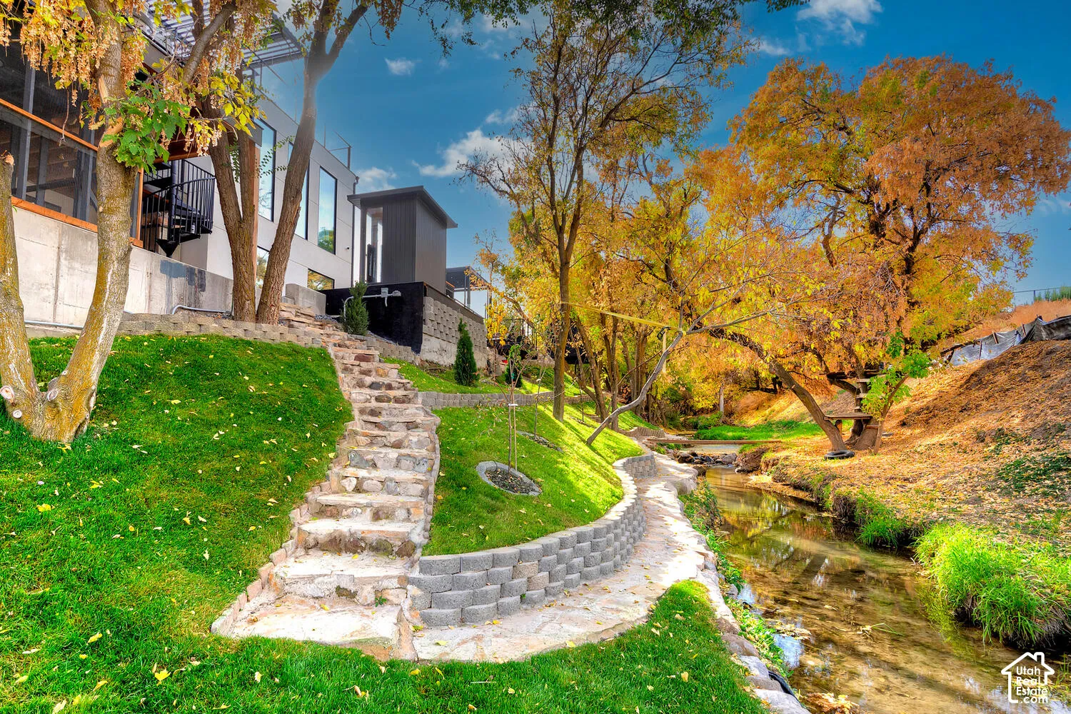 Exterior grounds featuring natural stone steps, segmented block retaining walls, mature trees, and a bordering stream