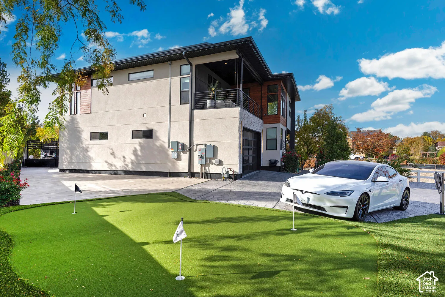 Contemporary exterior featuring textured stucco siding with wood-tone accents, an integrated upper-level balcony, a paved driveway, and a custom putting green