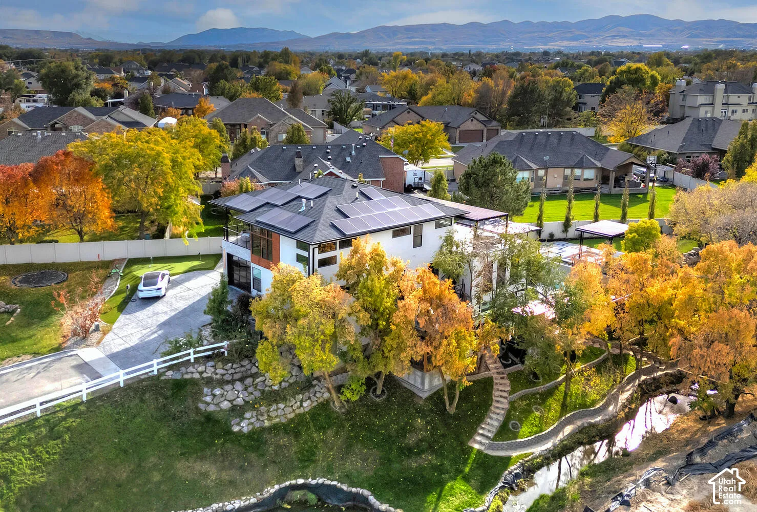 Aerial view of a residential property featuring roof-mounted solar panels, multi-level exterior decking, a stone-lined water feature, and a concrete driveway, set against a mountainous backdrop