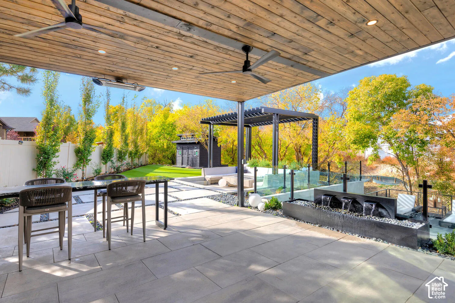 Covered patio featuring a wood-plank ceiling with recessed lighting and ceiling fans, stone paver flooring, a linear water feature with stone accents, a detached pergola, and a synthetic turf lawn
