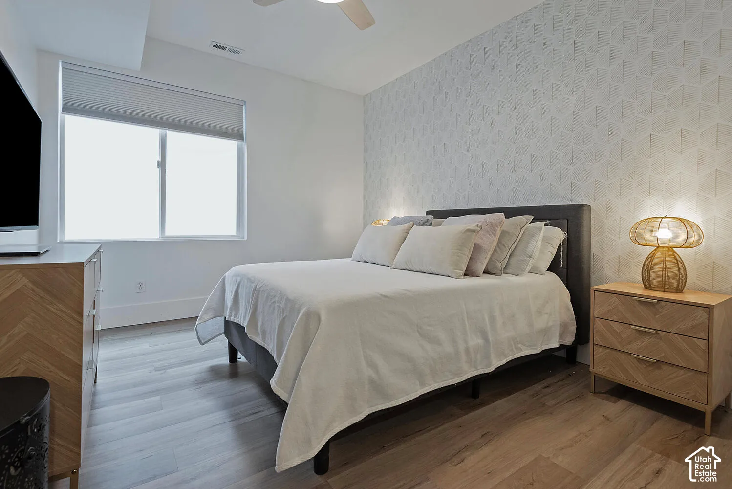 Room featuring wood-finish flooring, a textured geometric accent wall, a single-hung window with a neutral shade, and an integrated ceiling fan