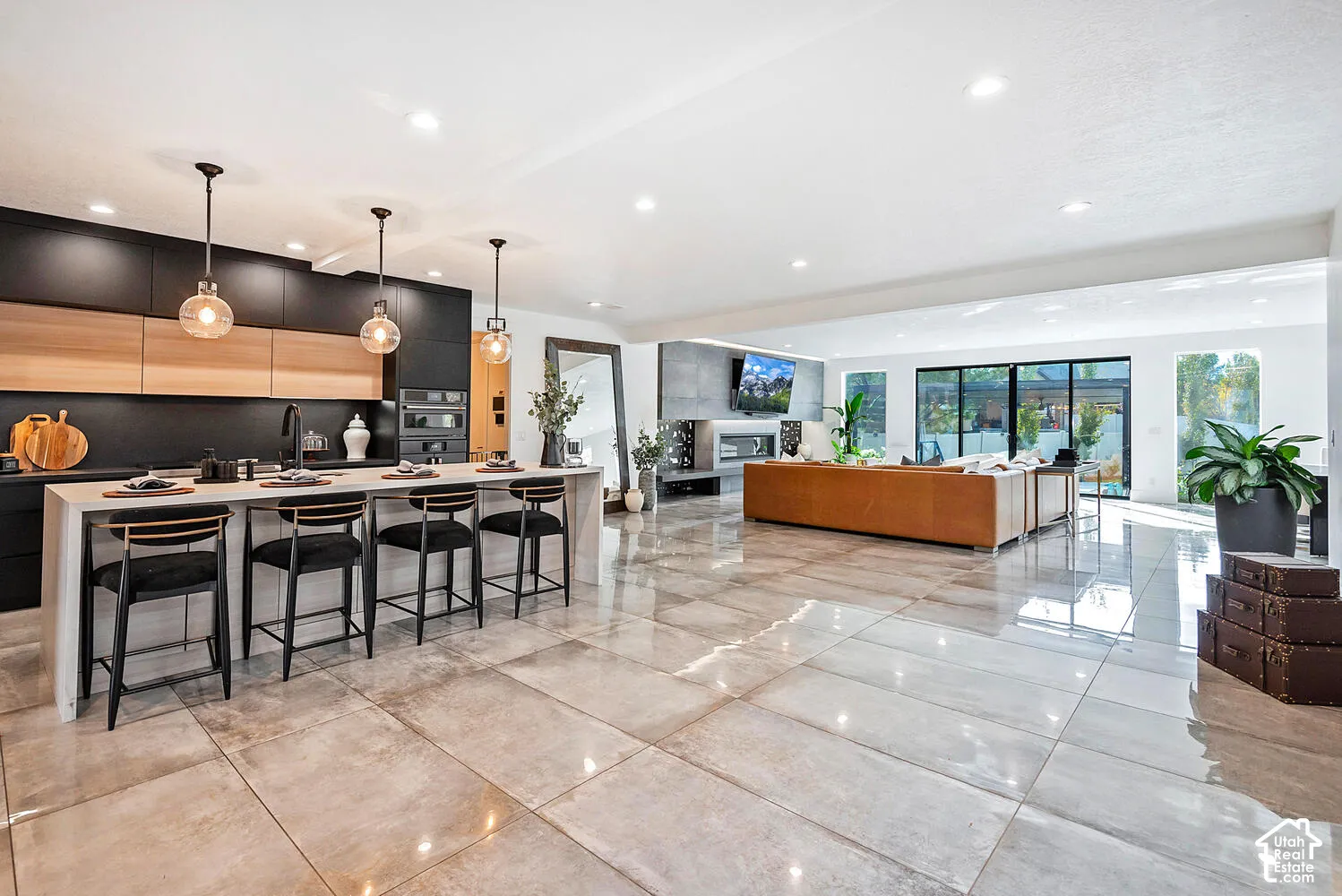 Open-concept kitchen and living area featuring oversized tile flooring, matte black cabinetry with natural wood accents, a waterfall-edge kitchen island, and a linear fireplace with stone surround