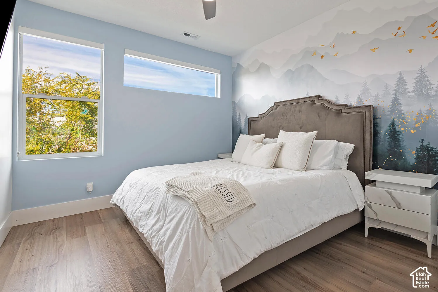 Bedroom featuring wood-finish flooring, a wall-length mountain and forest motif mural, double-pane picture windows, horizontal transom window, and a ceiling-mounted fan