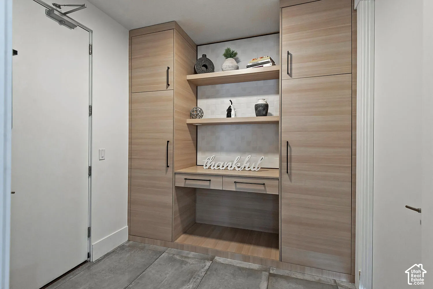 Built-in wood-finish cabinetry featuring dual full-height closets, two floating shelves, a desk surface with two drawers, and a tiled backsplash