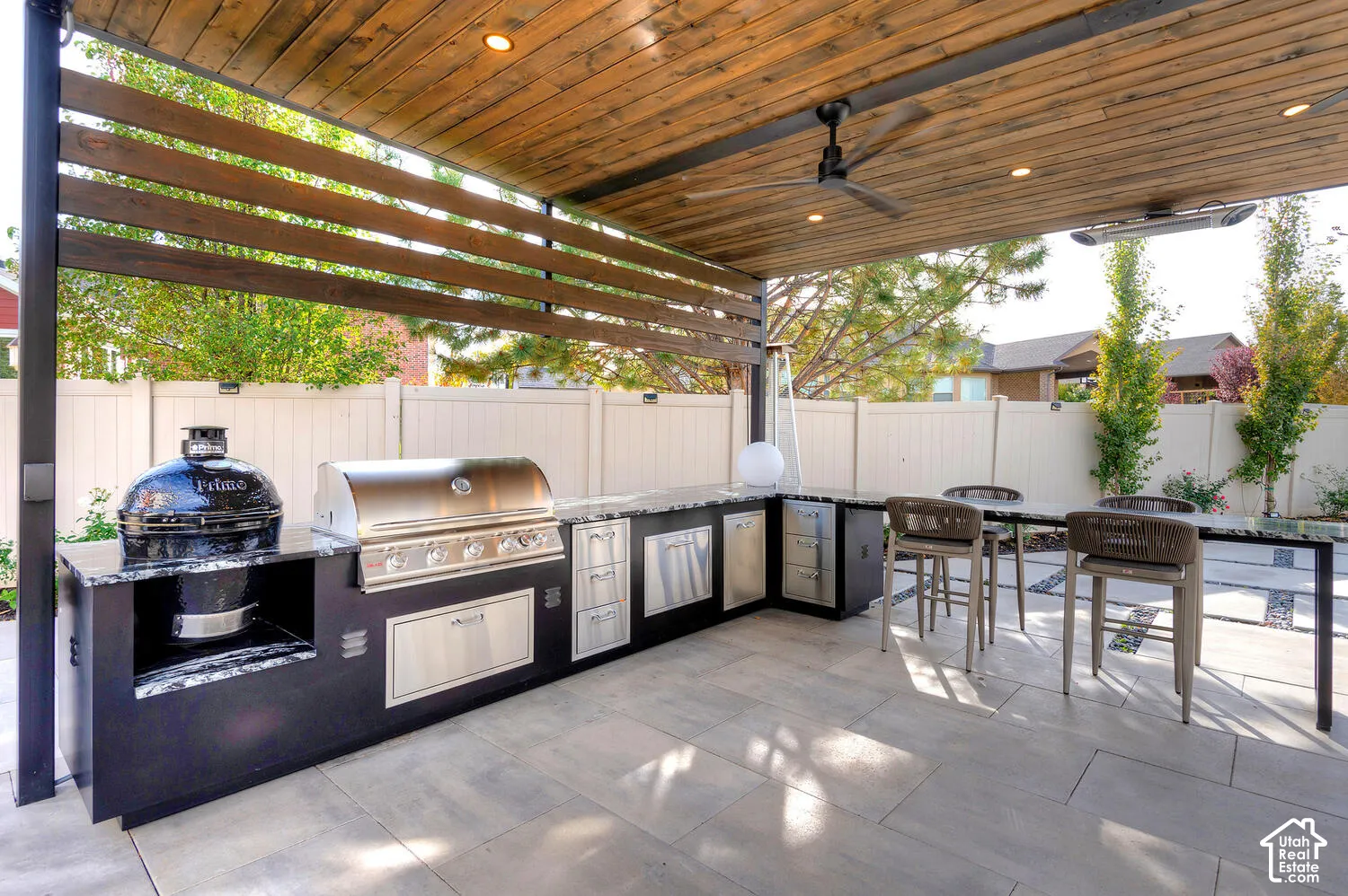 Covered outdoor kitchen featuring stone-finish countertops, stainless steel built-in grill and cabinetry, wood-slat ceiling with integrated lighting and ceiling fan, and large-format paver flooring