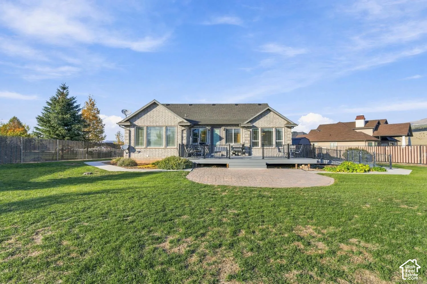 Rear exterior featuring a brick facade, raised deck with black metal railing, concrete paver patio, and expansive lawn