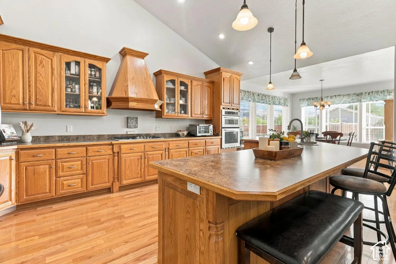 Kitchen featuring oak cabinetry, a custom range hood, double wall ovens, a large island with countertop seating, and vaulted ceilings with pendant lighting