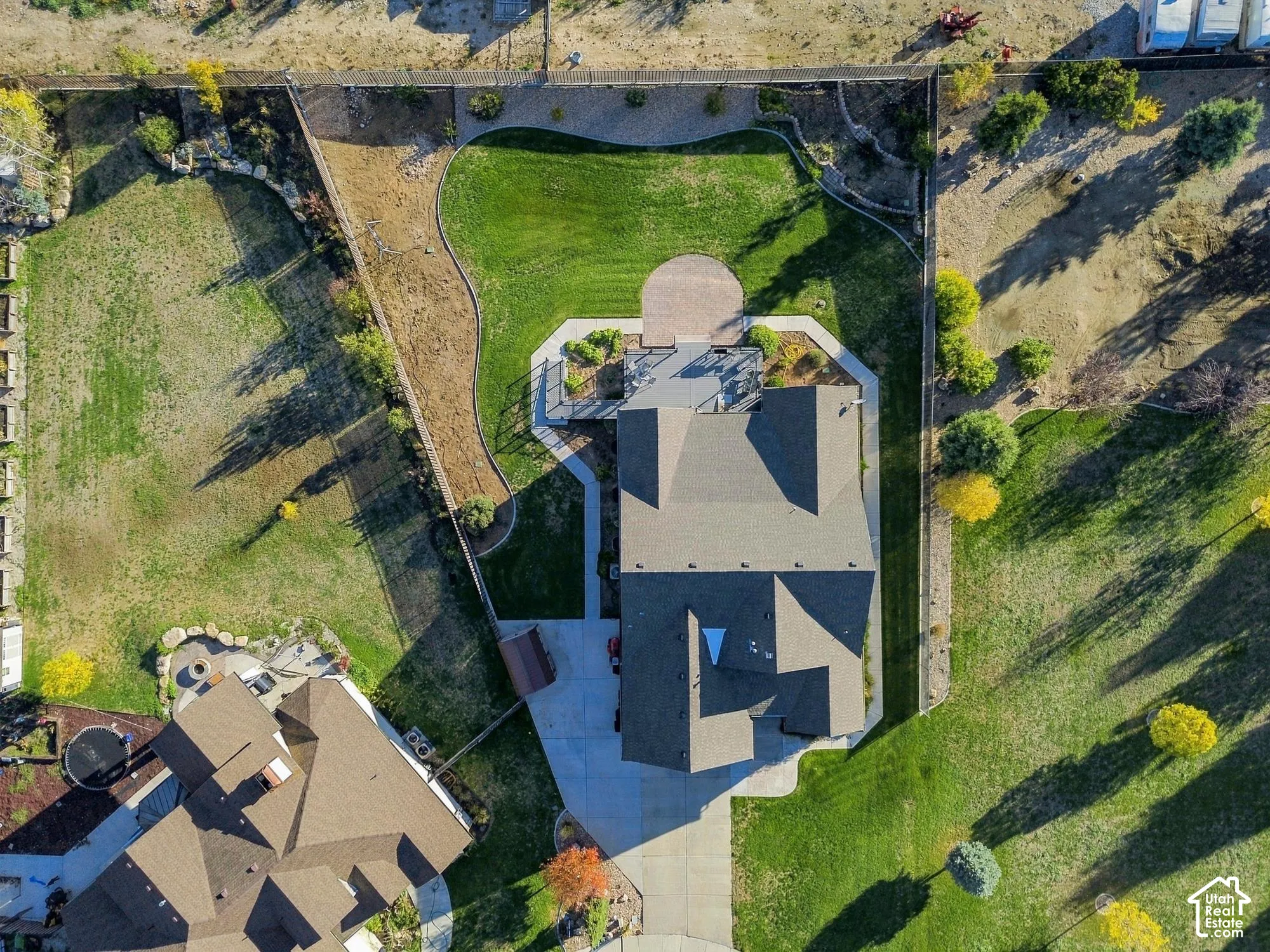 Aerial perspective of a sprawling corner lot featuring a manicured lawn, stone retaining walls, an expansive rear deck, a paved stone patio, and a concrete driveway