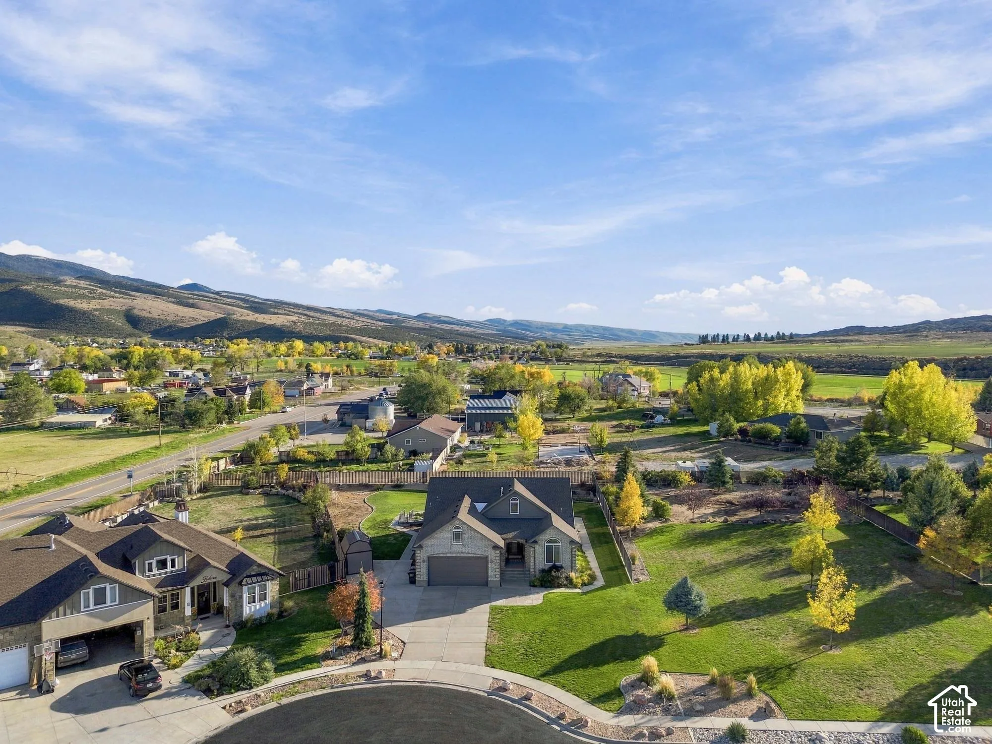 Aerial perspective of residential properties featuring expansive grass lots, mature trees, concrete driveways, and mountain scenery
