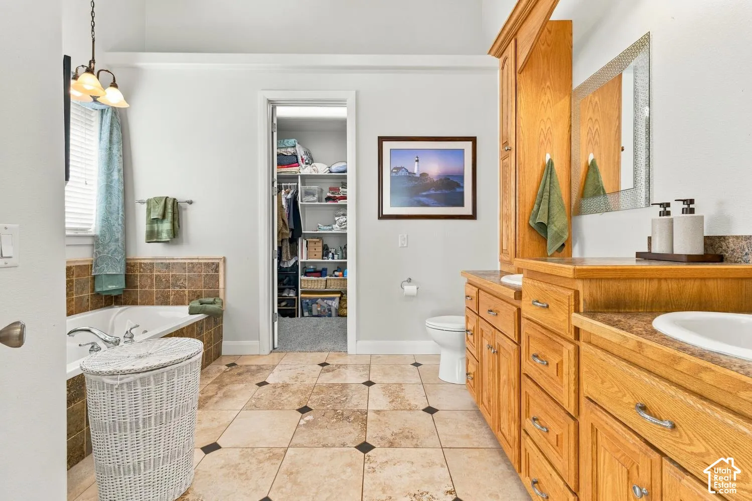 Bathroom featuring stone tile flooring with decorative inlays, dual-sink vanity with light wood cabinetry, tiled soaking tub surround, and a walk-in closet with built-in shelving