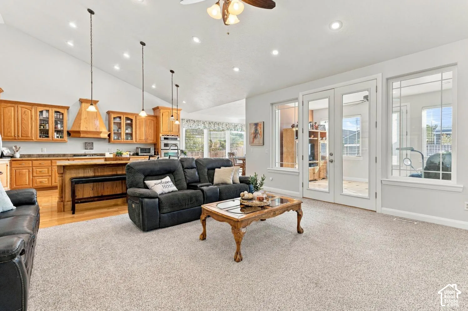 Open-concept floor plan featuring vaulted ceilings, recessed lighting, wood cabinetry with integrated range hood, a central kitchen island, and glass French doors