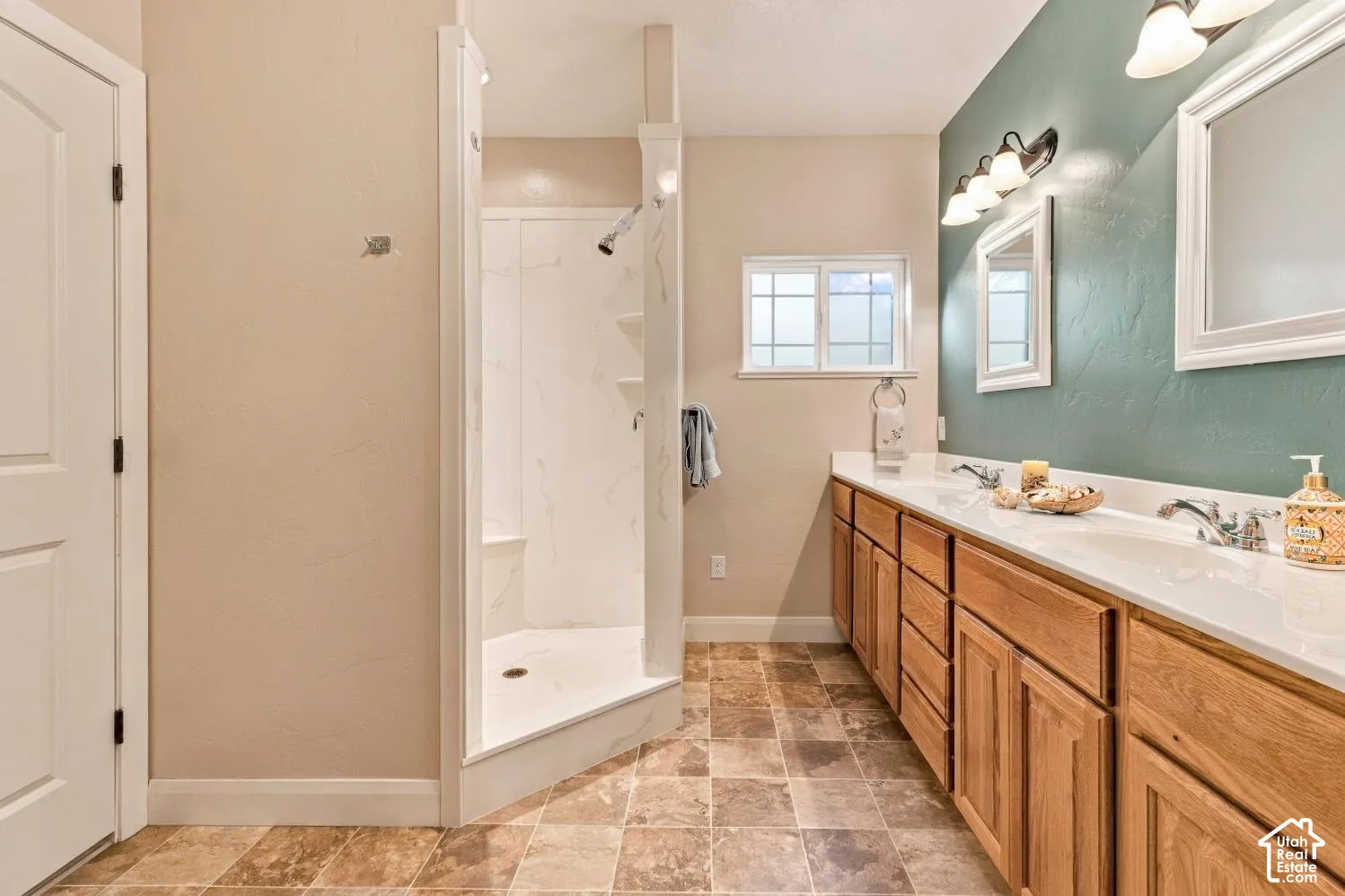 Bathroom featuring a double-sink vanity with wood cabinetry, a walk-in shower with integrated shelving, tile flooring, and wall-mounted light fixtures