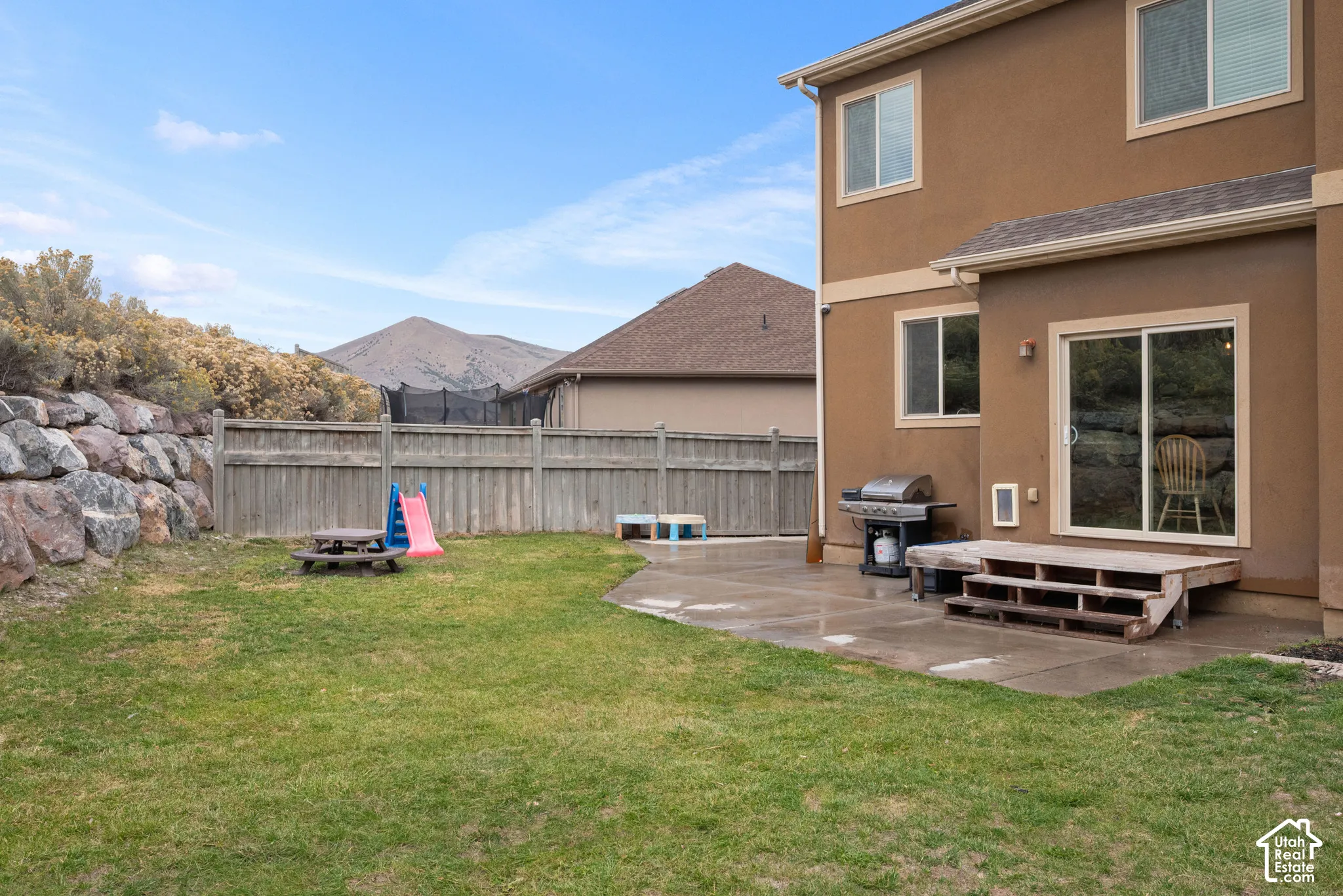 Rear exterior featuring a concrete patio, wood-frame staircase, rock retaining wall, and mountain backdrop
