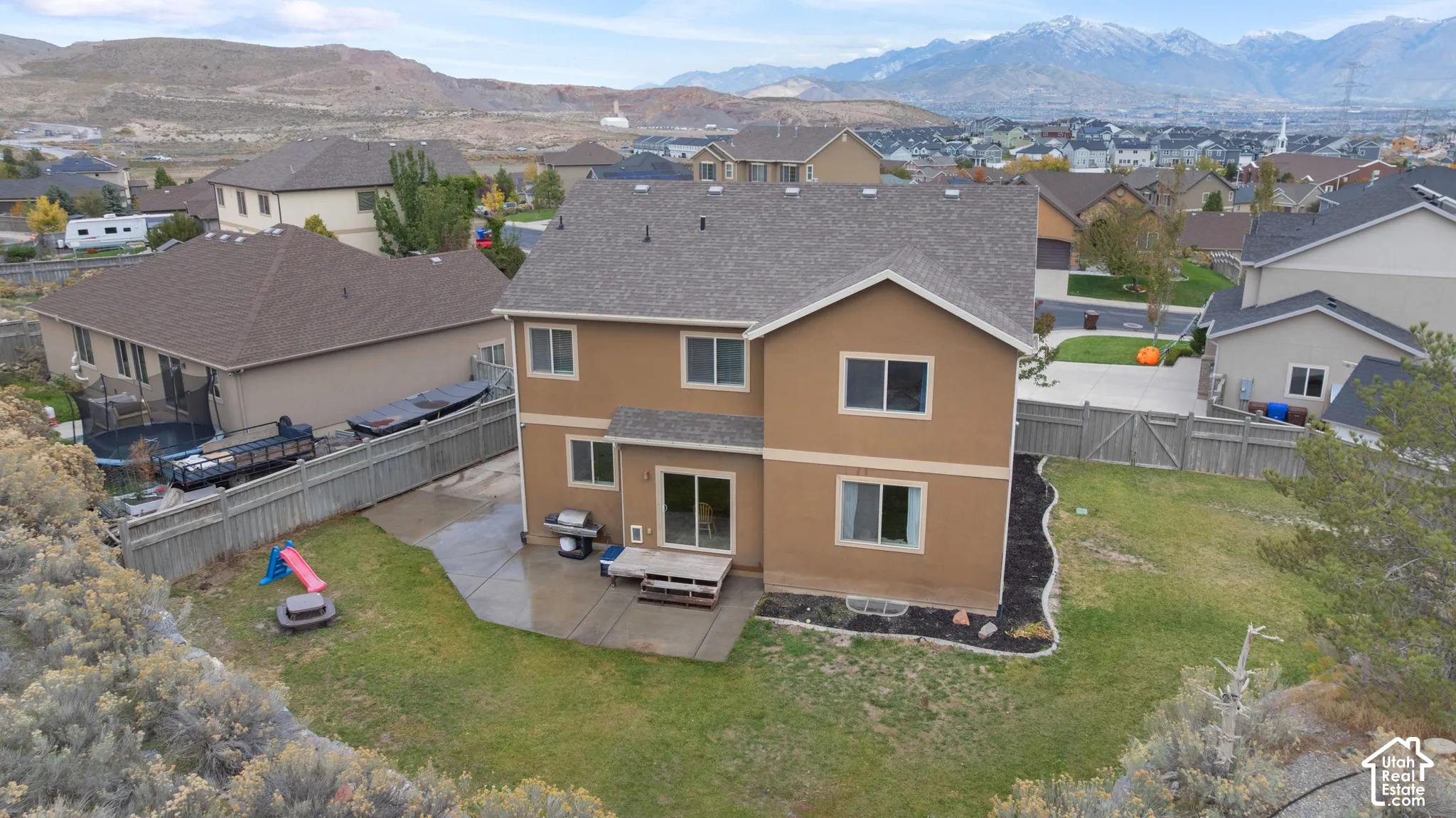 Two-story exterior featuring a stucco facade, asphalt shingle roof, concrete patio, perimeter wood fencing, and mountain backdrop