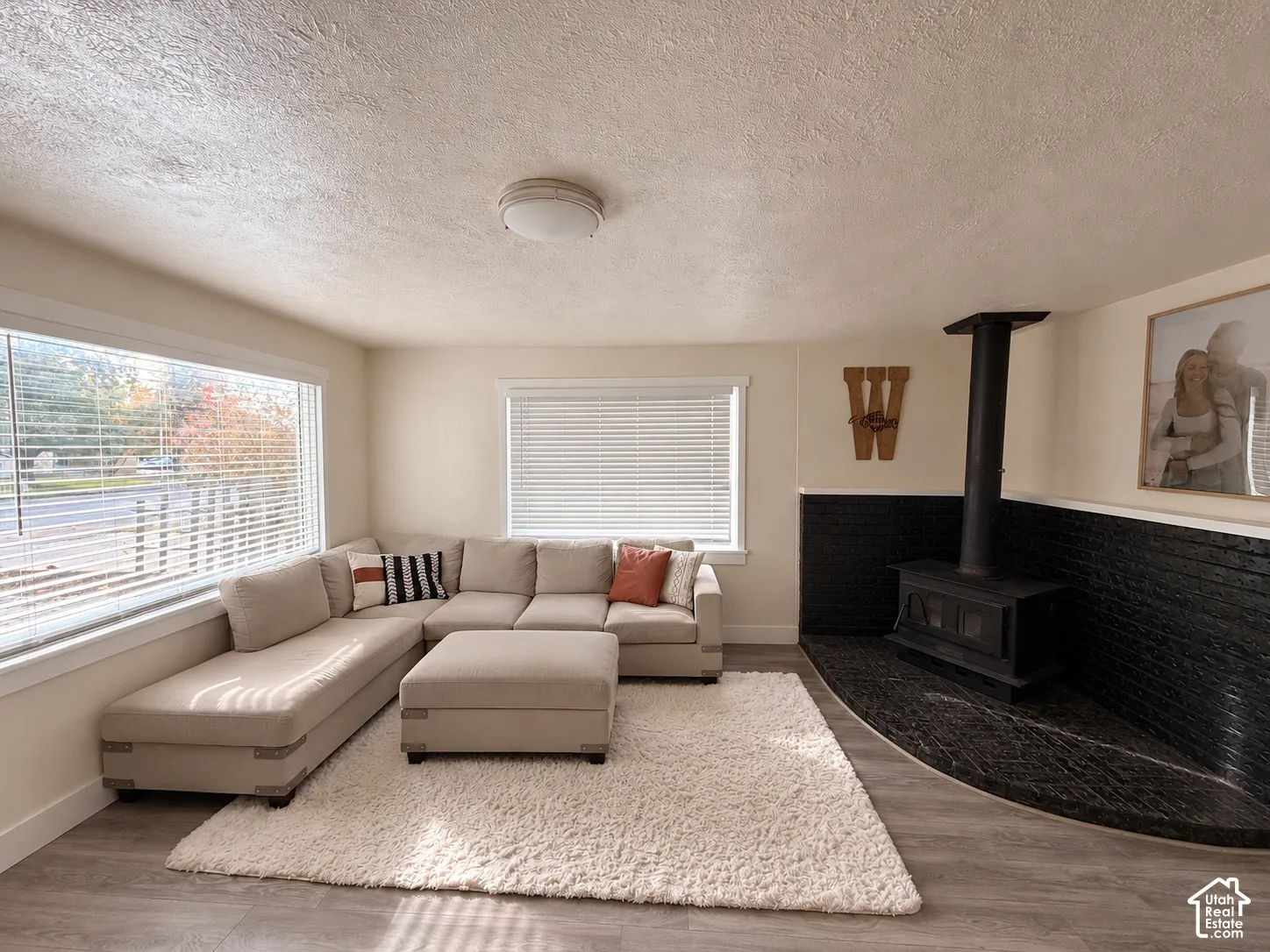 Living space featuring wood-finish flooring, large windows with horizontal blinds, and a wood-burning stove set against a black-painted masonry accent wall with a curved stone hearth