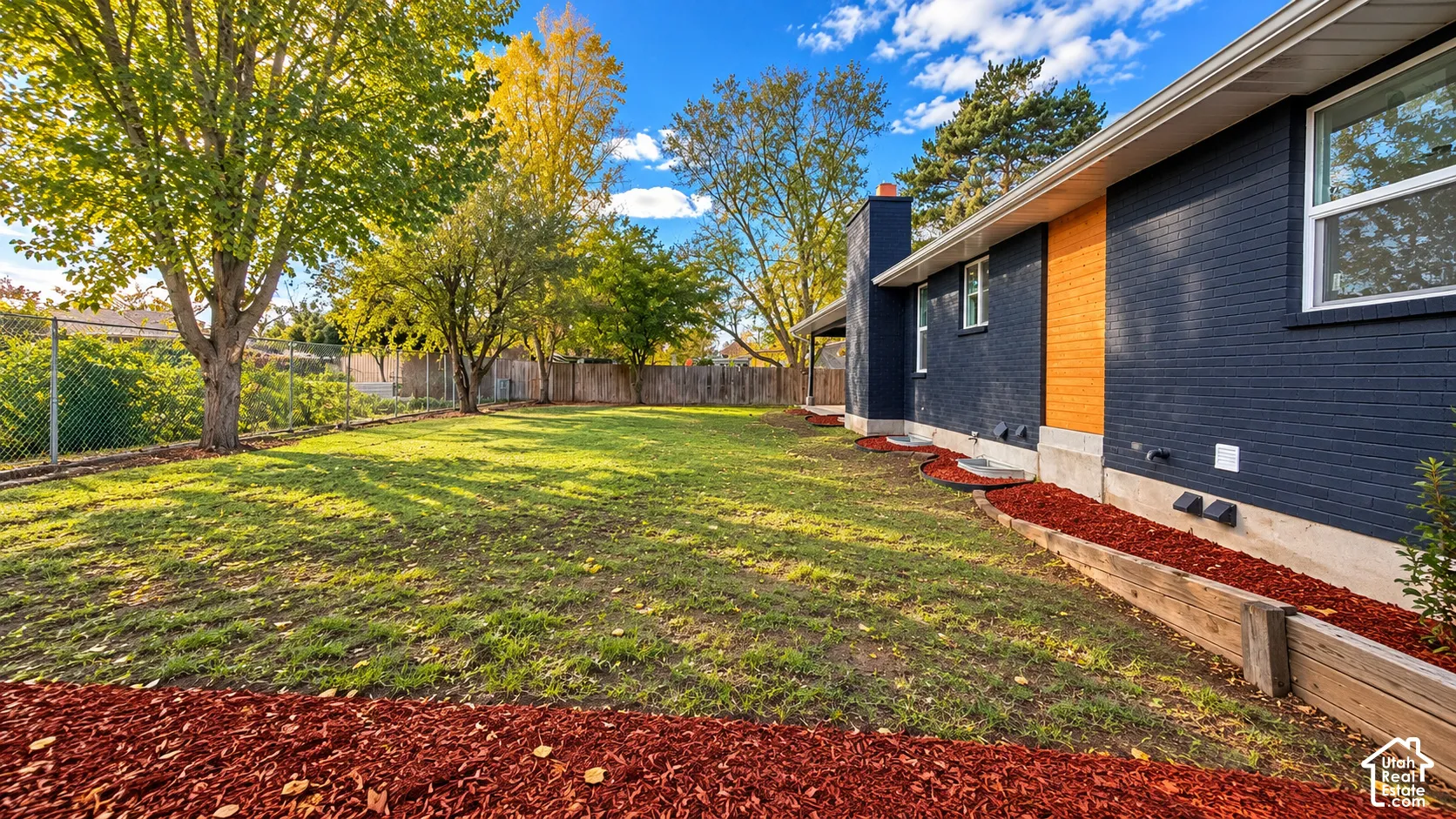 Expansive backyard featuring a manicured lawn, wood-trimmed garden beds, mature trees, and an exterior highlighted by dark-painted brick and wood-panel accents