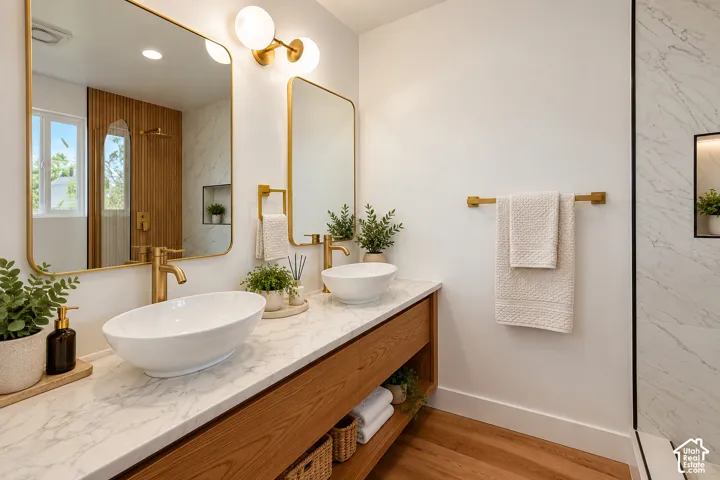 Double vanity featuring wood-finish cabinetry with an open shelf, stone-patterned countertop, oval vessel sinks, brass-tone hardware, and gold-framed mirrors