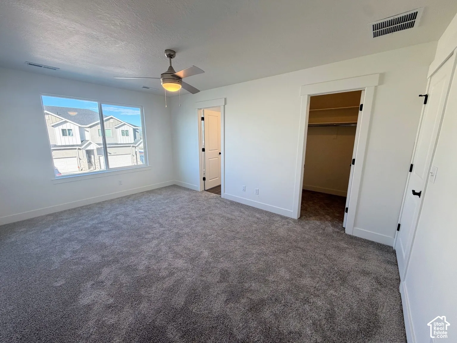 Neutral-toned wall-to-wall carpeting, ceiling fan with integrated lighting, oversized window, and walk-in closet with built-in shelving